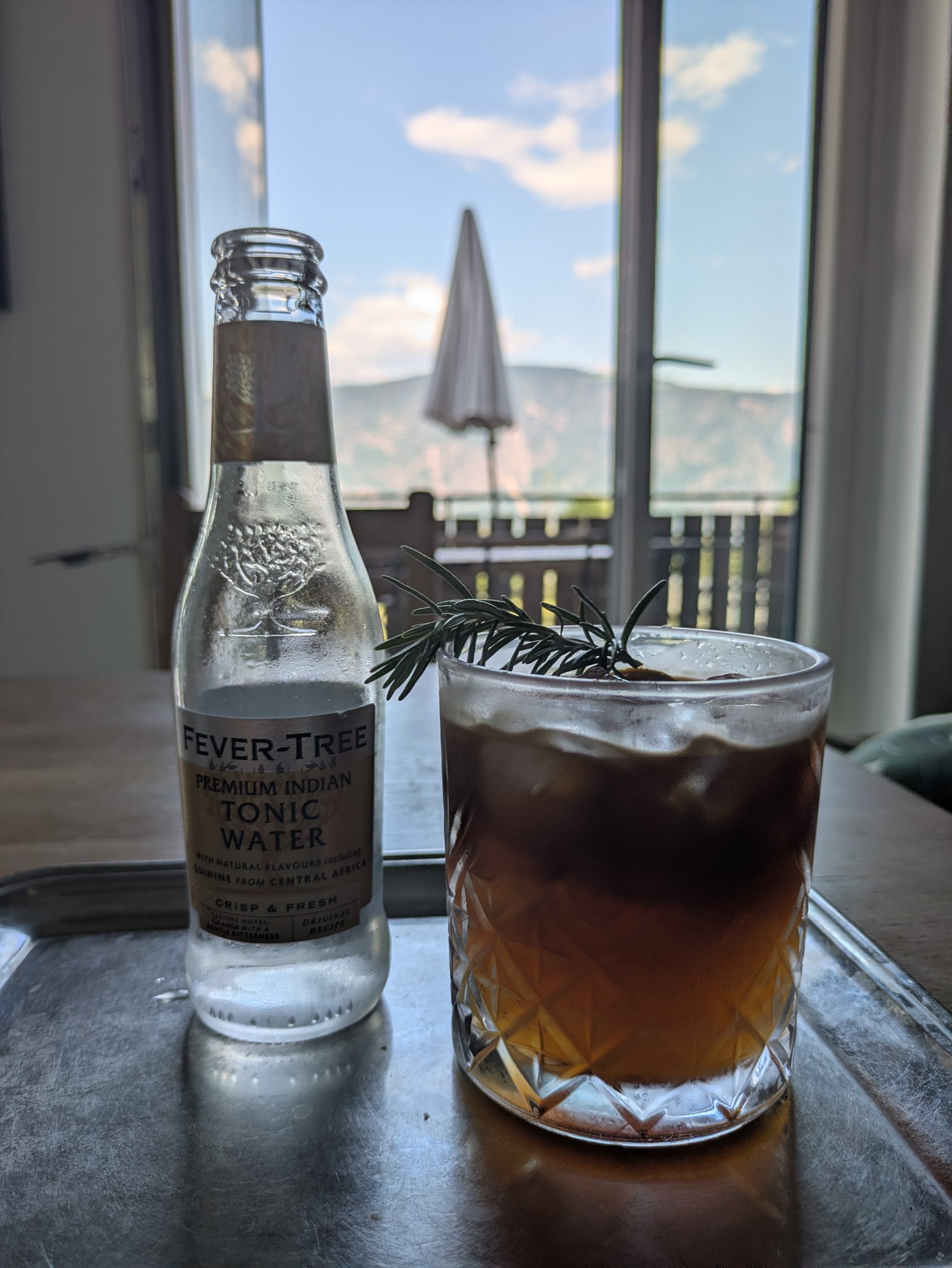 Ein Glas Espresso-Tonic mit Rosmarin-Garnitur auf einem Tisch, im Hintergrund der Balkon